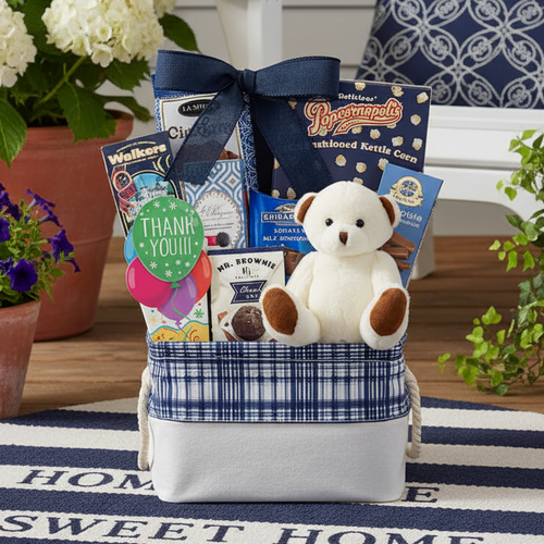 Gift basket with teddy bear, snacks, and a plush toy on a 'Home Sweet Home' rug with potted plants and a chair in the background.