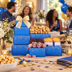 A tiered gift box arrangement filled with assorted treats, including popcorn, chocolates, and muffins, wrapped in bright blue paper with gold confetti and a large gold bow. The setup is on a decorated party table with balloons, flowers, and blurred people in the background.