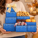 A person in a cream knit sweater holds a gift box arrangement made of blue boxes with gold speckles and a large gold bow. The box contains caramel popcorn, chocolate brownies, yellow muffins, and wrapped candies.