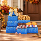 A holiday gift basket arrangement of blue gift boxes with white polka dots, filled with assorted treats including popcorn, cookies, chocolates, and candies, set on a wooden porch in front of a decorated door.