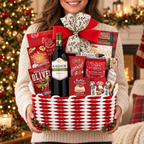 A woman in a cream knit sweater holds a large red-and-white woven gift basket filled with holiday treats and a bottle of FlakeMORE red wine. The basket is tied with a red bow and decorated with holly, set against a warm, blurred Christmas tree and fireplace background.