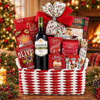 A holiday gift basket filled with assorted gourmet treats and a bottle of wine, tied with a red velvet bow and decorated with a red-and-white woven basket. Visible items include a bottle of Blakemore Cabernet Sauvignon, Olives by the Yard, Cookie Gallery, Bella Formaggio, and other festive snacks, set against a warm Christmas tree and fireplace background.