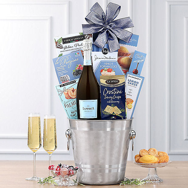 Gift basket with champagne, snacks, and pastries on a wooden table.